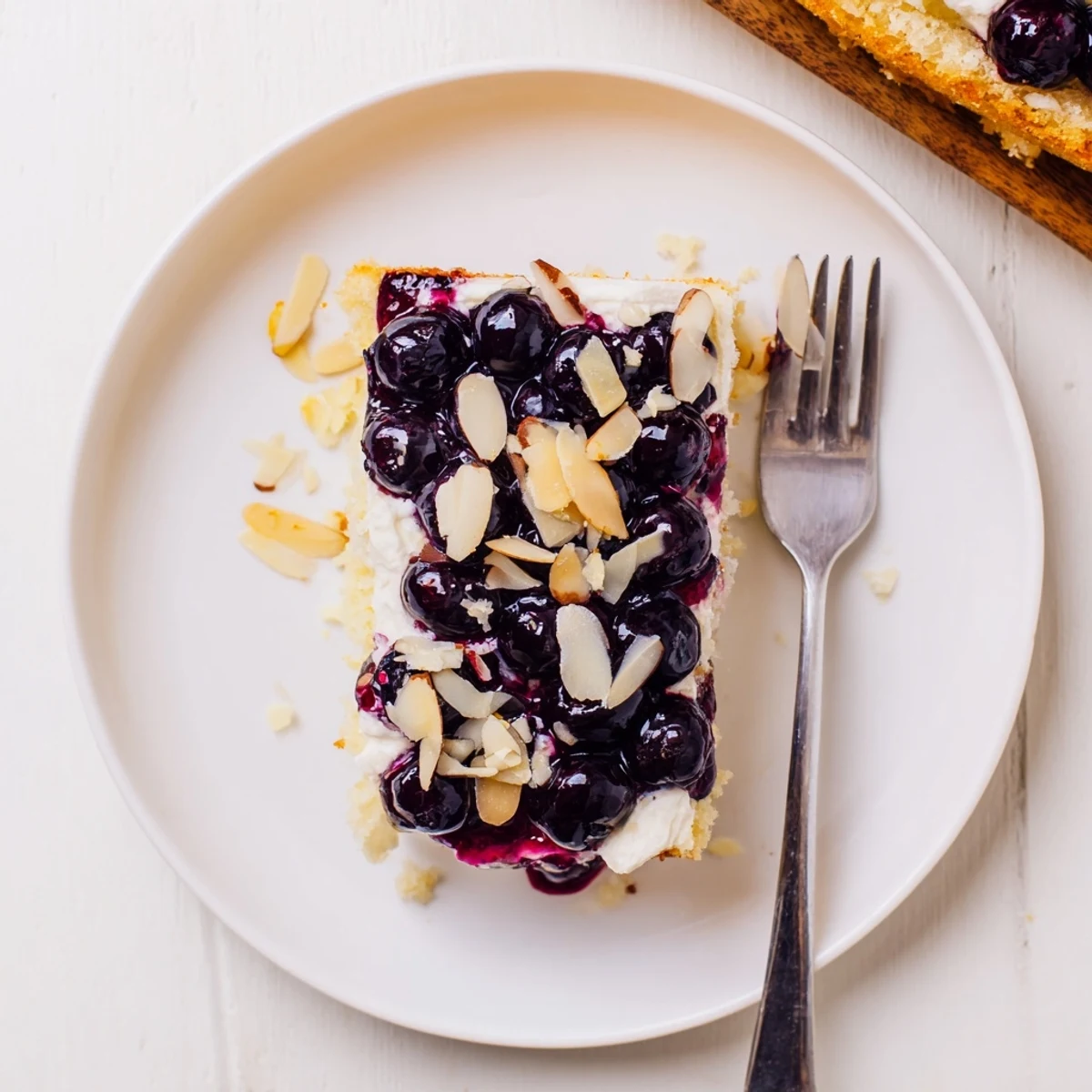 Zesty, creamy dessert slice shows Lemon Blueberry Poke Cake With Almond Topping served chilled.